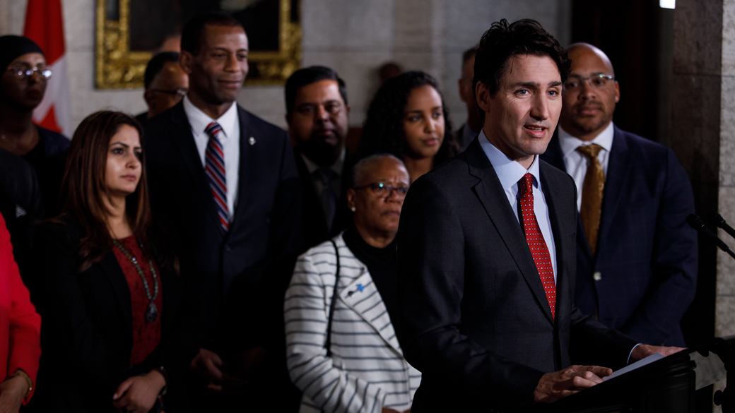 À l’intérieur du Parlement, des dirigeants de la communauté et des députés se tiennent derrière le premier ministre Trudeau pendant qu’il fait une annonce