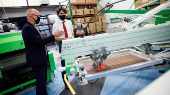 A man gestures with his hand near machinery, PM Trudeau and Minister Fortier look on