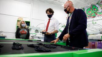 PM Trudeau holds a mask, a man stands at a machine with multiple masks, Minister Fortier looks on