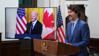 PM Trudeau smiles at a podium with a screen of President Biden smiling at a podium on screen
