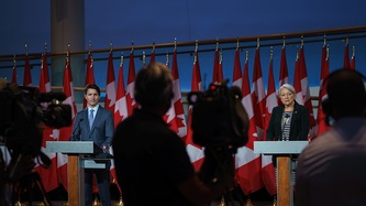 Prime Minister Justin Trudeau and Ms. Mary Simon stand at podiums facing camera technicians