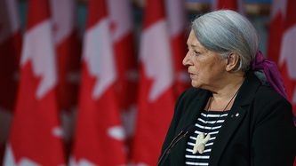 Ms. Mary Simon stands at a microphone with rows of Canadian flags in the background